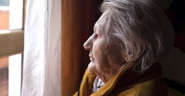 An older woman looking through a window from her living room
